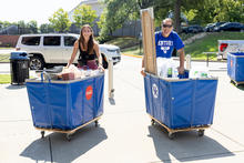 A smiling young woman and a man in a Kentucky Dad T-shirt push blue carts loaded with belongings on a sunny campus.