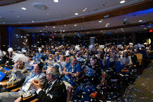 A large group of people seated in an auditorium, wearing blue and white attire and accessories such as scarves and pom-poms. Blue and white confetti fills the air, creating a festive atmosphere. 