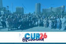 A large group of people pose together outdoors in front of several tall buildings on a sunny day. Streamers or ribbons can be seen in the sky above them. The lower portion of the image contains a white banner with the stylized text “NCUR 26 @ Richmond” in