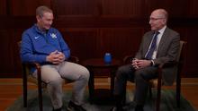 Two people sit across from each other in chairs on either side of a small round table in a wood‑paneled room. One wears a UK athletic-style jacket and light-colored pants, while the other wears a suit and tie. A small blue object sits on the table between