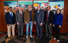 Six people stand side by side indoors, posing for a group photo in front of colorful artwork.