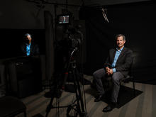 A person sits in a recording studio during a video interview. A camera, lighting equipment and microphones are visible in the foreground, capturing the subject against a dark backdrop while a monitor displays the live video feed.