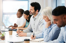 A group of people seated at a conference table, taking notes and working on laptops during a meeting or training session.
