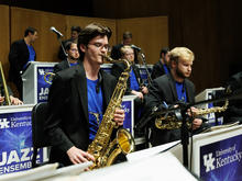 white male in a black jacket and blue shirt playing tenor saxophone in a jazz ensemble