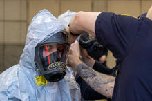 A responder in a protective hooded suit wears a full-face respirator while another responder adjusts the mask straps during an emergency preparedness drill.
