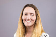 Portrait of a smiling woman with long blonde hair wearing a white polka-dot blouse against a light gray background.