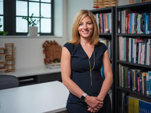 Person standing in a modern office, wearing a dark dress, with bookshelves in the background.