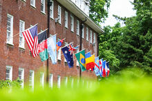 Row of international flags displayed on poles outside a red brick building, with green foliage in the foreground.