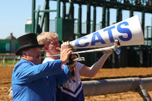 Two people at a horse racetrack, one playing a trumpet and one shouting into a megaphone labeled “CATS.”
