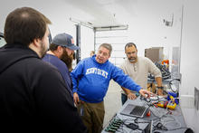 Group of people gathered around a workbench, looking at electronics and tools in a workshop.