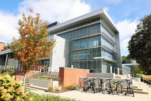 A modern, multi-story glass academic building sits on a landscaped campus, with a small tree showing autumn colors in the foreground and a bike rack filled with bicycles near the entrance on a sunny day.