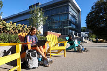 Student sitting in a bright yellow Adirondack chair outside a modern glass campus building, working on a laptop with a backpack and water bottle nearby, while other students sit and study along the walkway on a sunny day.