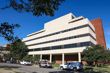 This is an exterior photo of the College of Nursing Building at UK, a multistory building with a white stone front lined with columns along a street with parked vehicles and a clear blue sky. 