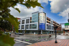 Modern multi-story academic building on a rain-slicked city corner, framed by green leaves, with pedestrians and light traffic at the intersection.