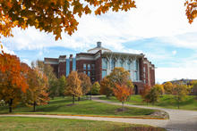 A large brick academic building sits on a gently sloping hill, surrounded by trees with bright autumn foliage in shades of orange, red, and yellow. A winding paved path curves through the grassy landscape under a partly cloudy sky.