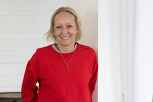 Vikki Lane smiles at the camera while standing in front of a white wall. She is wearing a red sweater with a gray collar and a silver necklace.