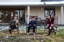 Three people sitting on wooden rocking chairs in a grassy yard with light snow, in front of a house with a covered porch.