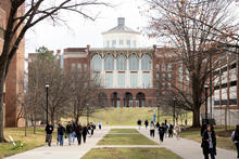 A wide campus walkway lined with leafless trees and grass leads toward a large academic building with arched windows and a central cupola, while groups of students walk along the path on an overcast day.