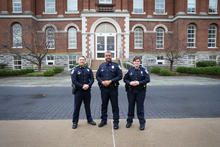 Three University of Kentucky Police Department officers in uniform stand on a campus walkway in front of a historic red-brick building with arched entry and stone base.