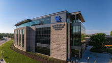 Aerial view of the University of Kentucky’s Martin-Gatton Agricultural Sciences building, featuring a stone-and-glass exterior and a large UK logo under a clear blue sky.