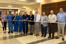A group of hospital staff and administrators stand in a lobby holding scissors and a ribbon during a ribbon‑cutting ceremony. 