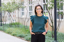 A person stands on a landscaped campus walkway outside a building, wearing glasses and a dark green blouse, with trees and greenery in the background.