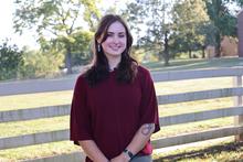 Person standing outdoors in front of a wooden fence, with trees and a grassy area in the background.
