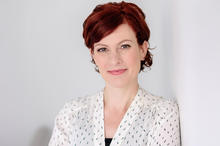 A person with short, wavy red hair stands against a plain light-colored wall, wearing a white blouse with a subtle dotted pattern.