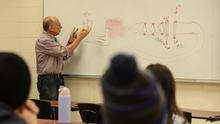A professor teaches in front of a classroom whiteboard covered with scientific diagrams and notes while students listen from their seats.