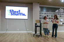 Two presenters stand at the front of a room giving a presentation beside a large screen displaying the logo “Sheet Shuffle.” One person speaks into a microphone while the other stands near a laptop on a cart. A photo wall is visible in the background.