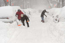 People shoveling snow on a street lined with cars during a heavy snowstorm.”