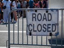 Metal barricades block a street with a white sign reading “ROAD CLOSED,” while pedestrians are visible in the background.