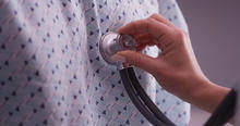 A health care provider places a stethoscope against the chest of a person wearing a hospital gown.