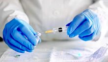 Close-up of blue-gloved hands holding a syringe filled with yellow liquid above a surface covered with papers.