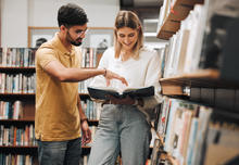 Two people looking at a book at a bookshelf. 