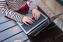 Person wearing a striped long‑sleeve shirt typing on a laptop at an outdoor wooden table.