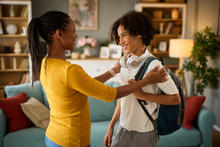 A young person wearing a backpack and headphones around their neck stands in a living room while another adult places their hands on the young person's shoulders. Bookshelves, a couch with pillows and a lamp are visible in the background.