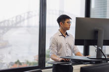 Person works at a standing desk in an office with floor-to-ceiling windows overlooking a harbor bridge and city skyline.