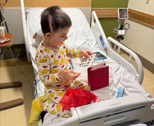A young child wearing patterned pajamas sits upright in a hospital bed, opening a small red gift bag filled with Valentine-themed items. Medical equipment is visible around the bed, and the child appears focused on exploring the contents of the bag.