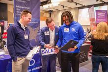 Three people stand at an event booth discussing information on a tablet, with a table displaying promotional materials and a visible “SALE” sign in the foreground, while other attendees browse nearby exhibits.