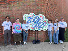 Six people stand on a sidewalk in front of a brick wall beside a large colorful sign reading “250 Lex.” Five wear white logo T-shirts, and one wears a bright tie-dye shirt.