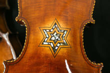 A close-up of the back of a violin featuring an inlaid six-pointed star design made of light-colored decorative materials. The wood has a warm, glossy brown finish with visible grain.