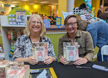 Two people sit at a table at an indoor event, smiling while holding up copies of a book. A display of books and posters is visible behind them.