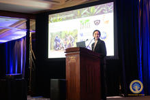 A person stands at a wooden podium presenting in a conference room. A large screen behind them displays a slide with photos of fieldwork and several logos. Blue curtains frame the stage, and an emblem is visible on the lower right side of the image.