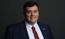 Studio portrait of a person in a navy suit, white shirt and red striped tie against a dark gradient background.