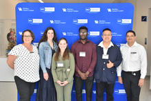 Six people pose in front of a blue step-and-repeat banner displaying the University of Kentucky Markey Cancer Center and NCI Comprehensive Cancer Center logos. Several individuals are wearing name badges.