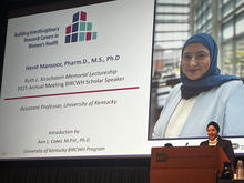 A presenter stands at a podium delivering a talk in front of a large projected slide. The slide shows a portrait and text about a research lecture on women’s health, including the speaker’s name, academic credentials, and affiliation with UK.