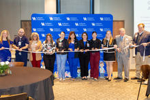 A group of people stand in a line holding a long ribbon for a ribbon‑cutting ceremony in front of a blue backdrop with the UK Golisano Children’s Hospital logo.