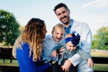 Two adults holding two young children outdoors near a wooden fence with green trees and a clear sky in the background.