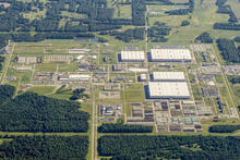 Aerial view of a large industrial complex surrounded by dense forest and open fields. The facility includes multiple large white-roofed warehouse-style buildings, grid-like roadways, smaller structures, parking areas, and neatly arranged outdoor storage.